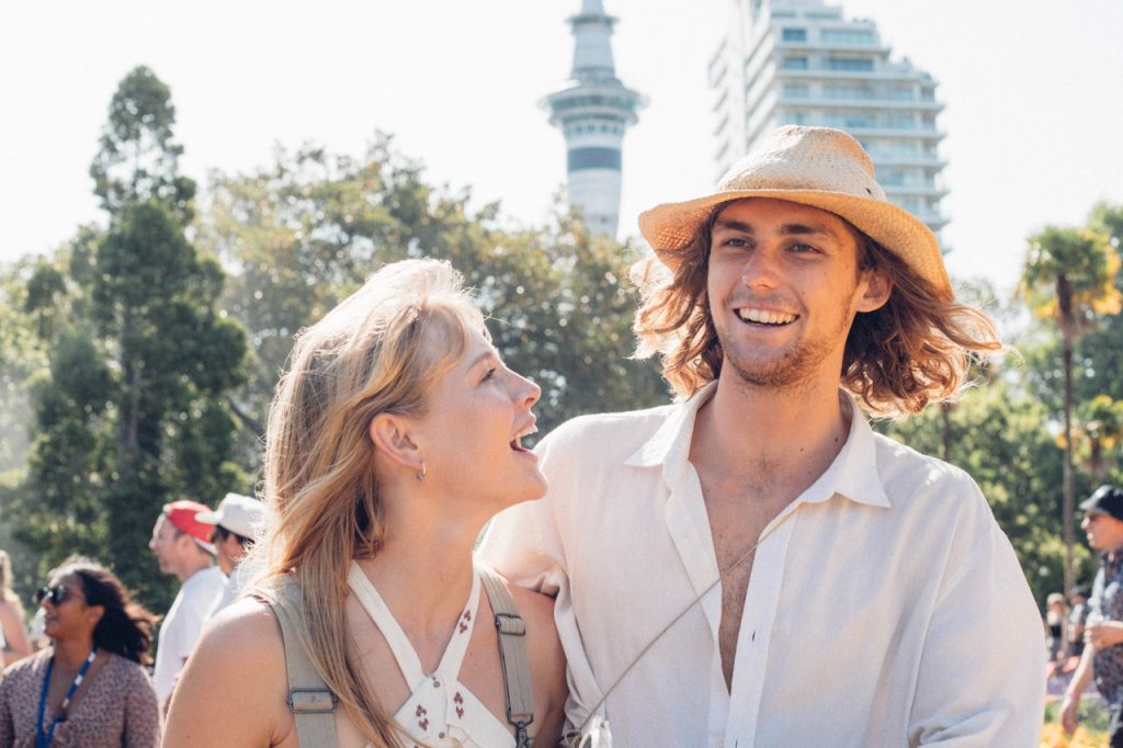 A-couple-laughing-in-a-city-park-with-people-the-Auckland-Sky-Tower-and-a-high-rise-building-behind-them